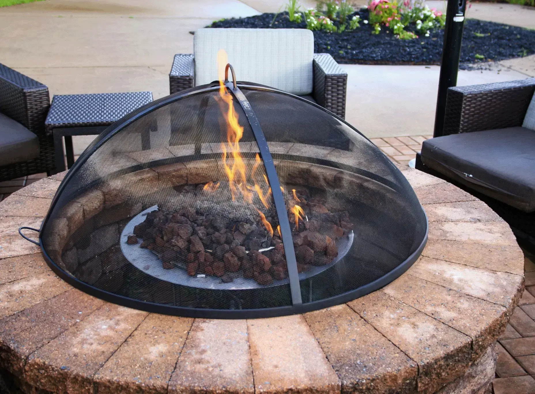 Fire pit with flames, surrounded by brick, with outdoor furniture in the background.