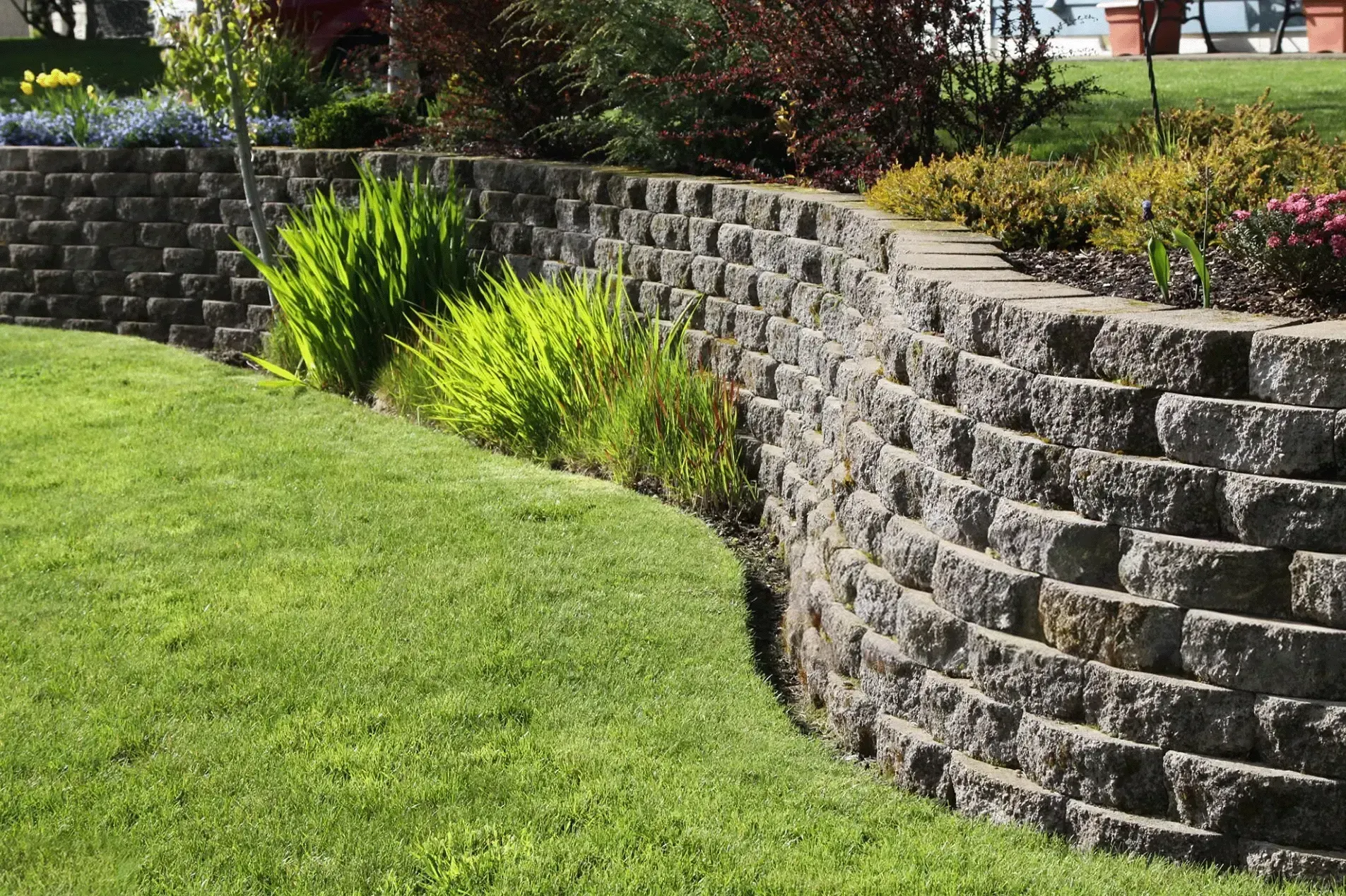 Curving gray brick retaining wall in garden with grass and greenery.