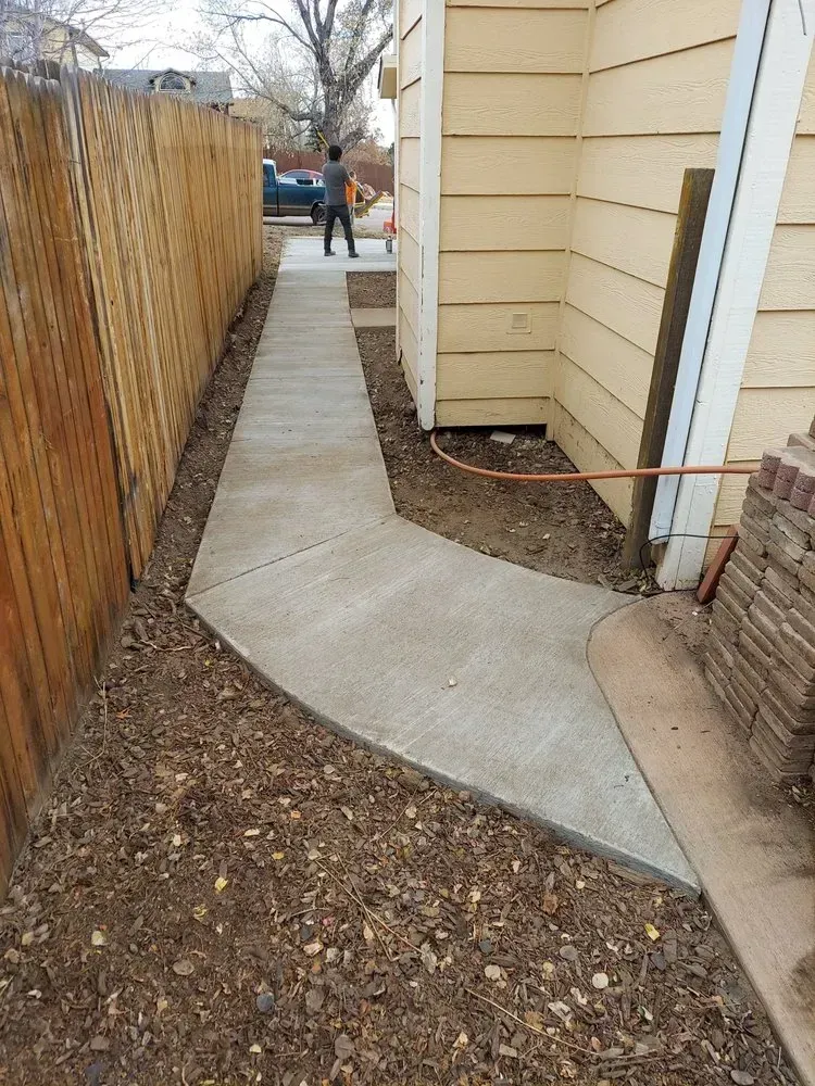 Concrete walkway between a wooden fence and a yellow house, person walking at the end.