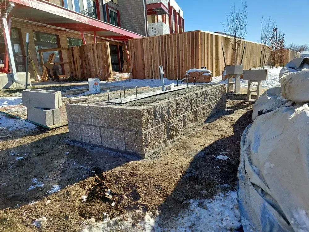 Construction site with cinder block structure, wooden fence, and building. Snowy ground, daylight.