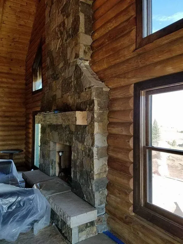 Stone fireplace in a log cabin setting with wood paneling and a window on the right.