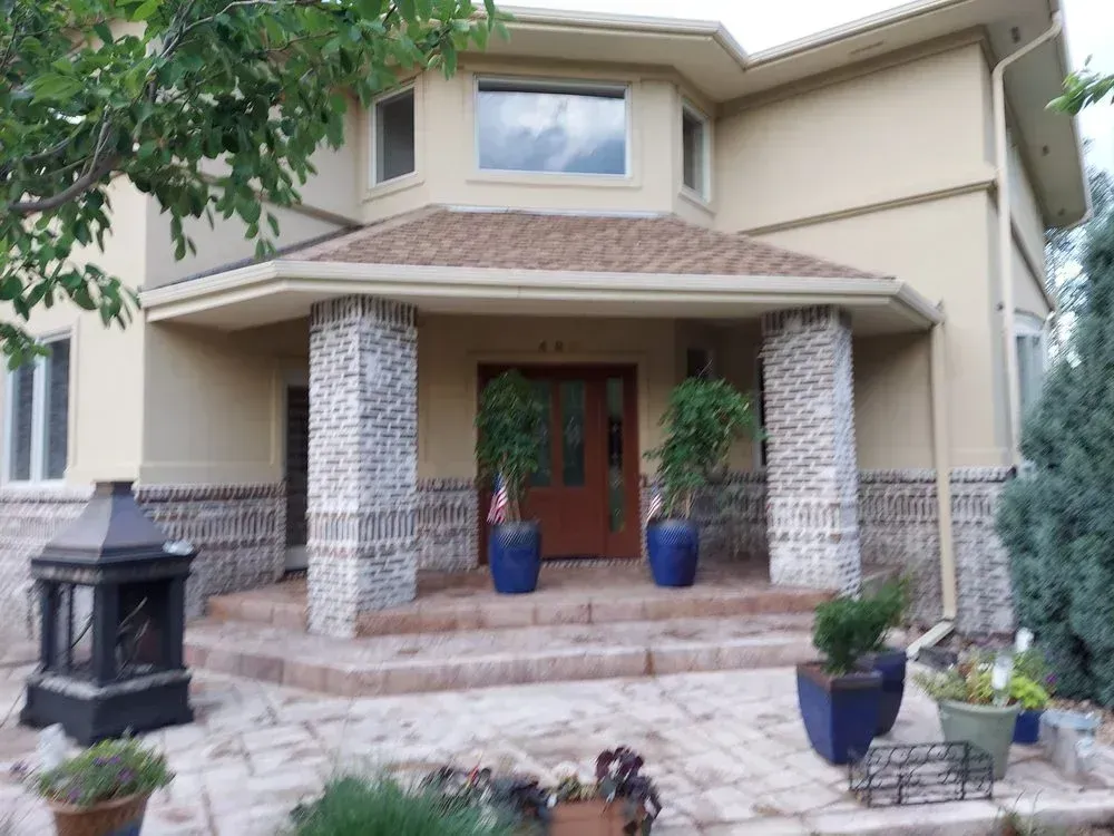 Beige two-story house with brick pillars, brown door, and plants in front.