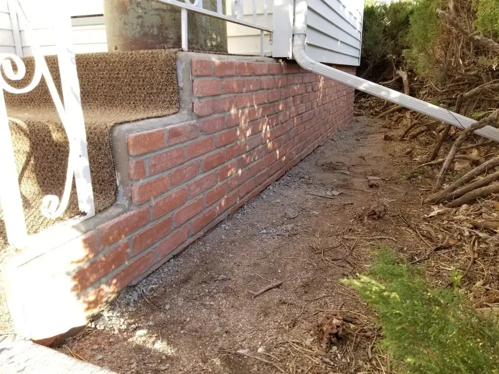 Brick steps leading up to a white house, surrounded by dirt and shrubbery.