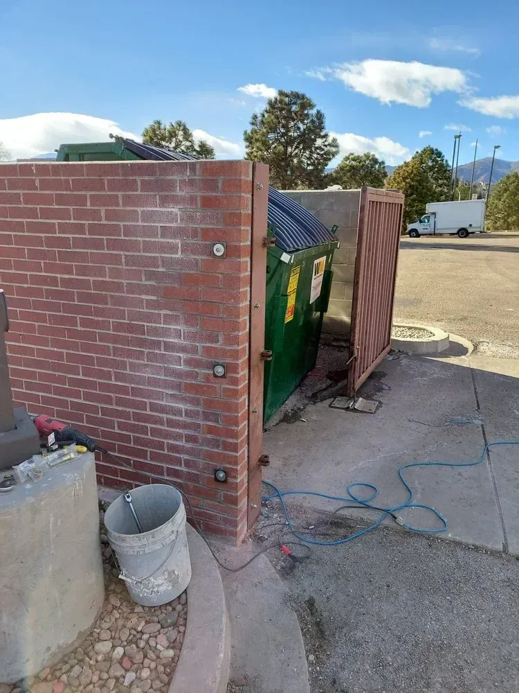 Brick enclosure around a green dumpster. Ajar gate, blue hose, sunny outdoor setting.