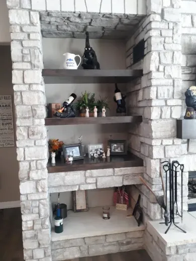 Stone-surrounded shelves with decor: wine bottles, plants, photos, and a fireplace in a living room.