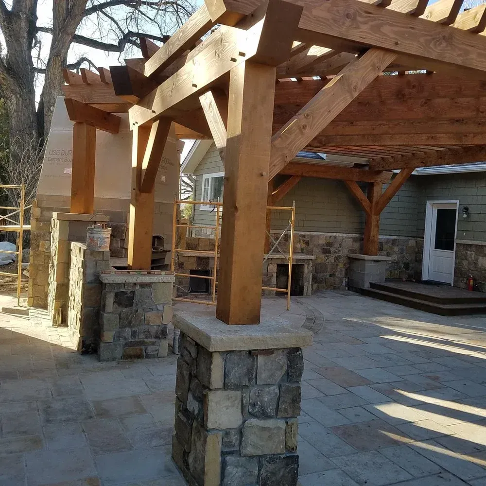 Wooden pergola over stone patio with stone pillars, next to a house.