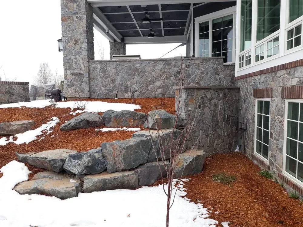 Stone steps and retaining walls next to a house with a covered patio, some snow, and mulch.