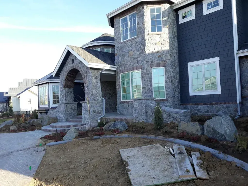 Stone and gray-sided house with curved entrance, multiple windows, and landscaping.