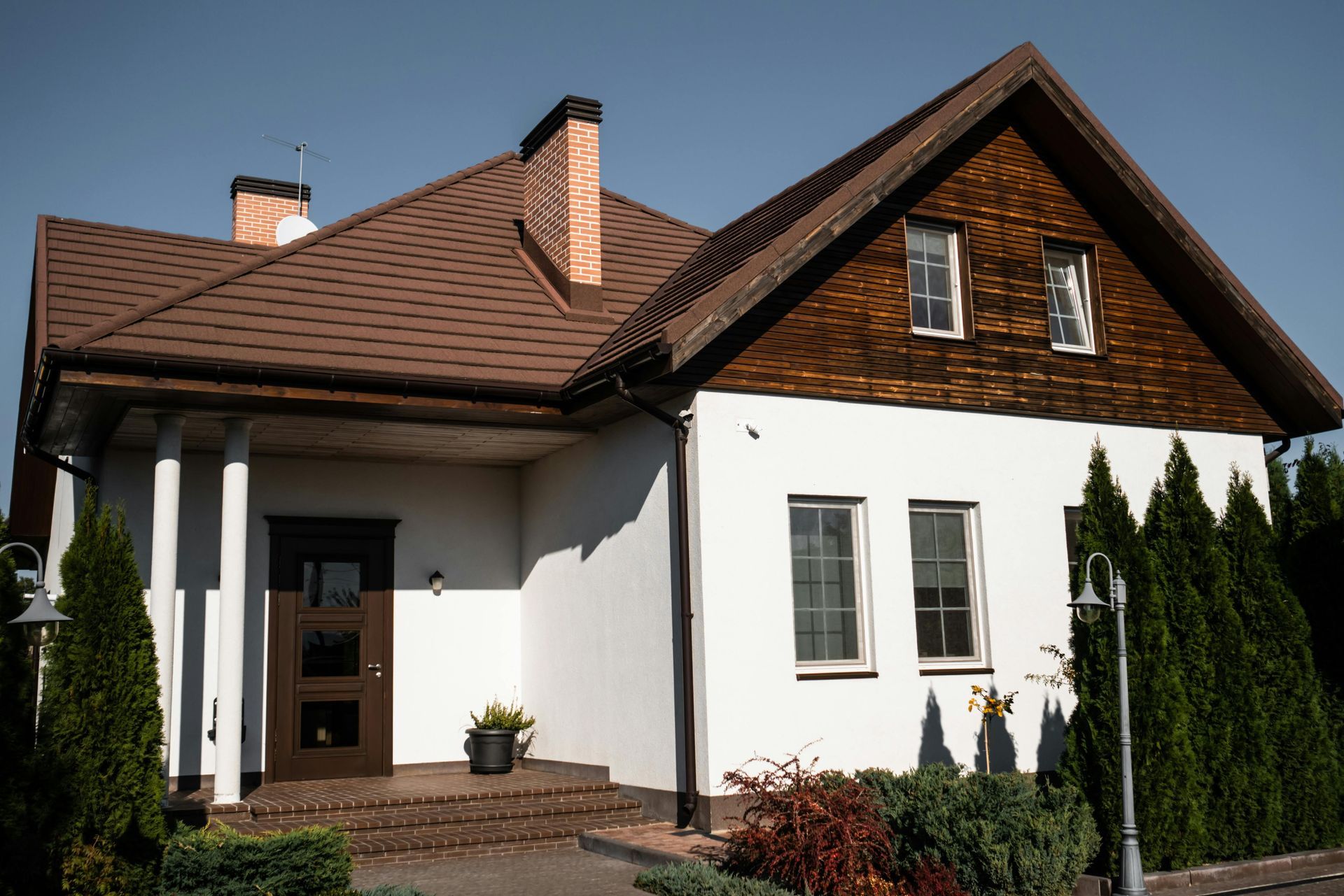 White house with brown roof and door, front porch with columns, trees and bushes.