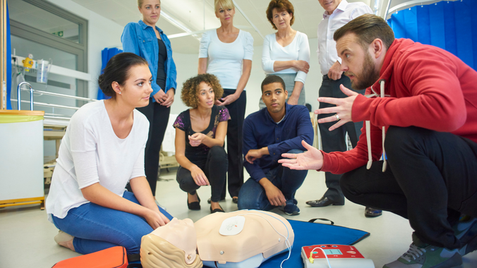 A group of people are sitting around a mannequin and a man is teaching them how to do a heart massage.