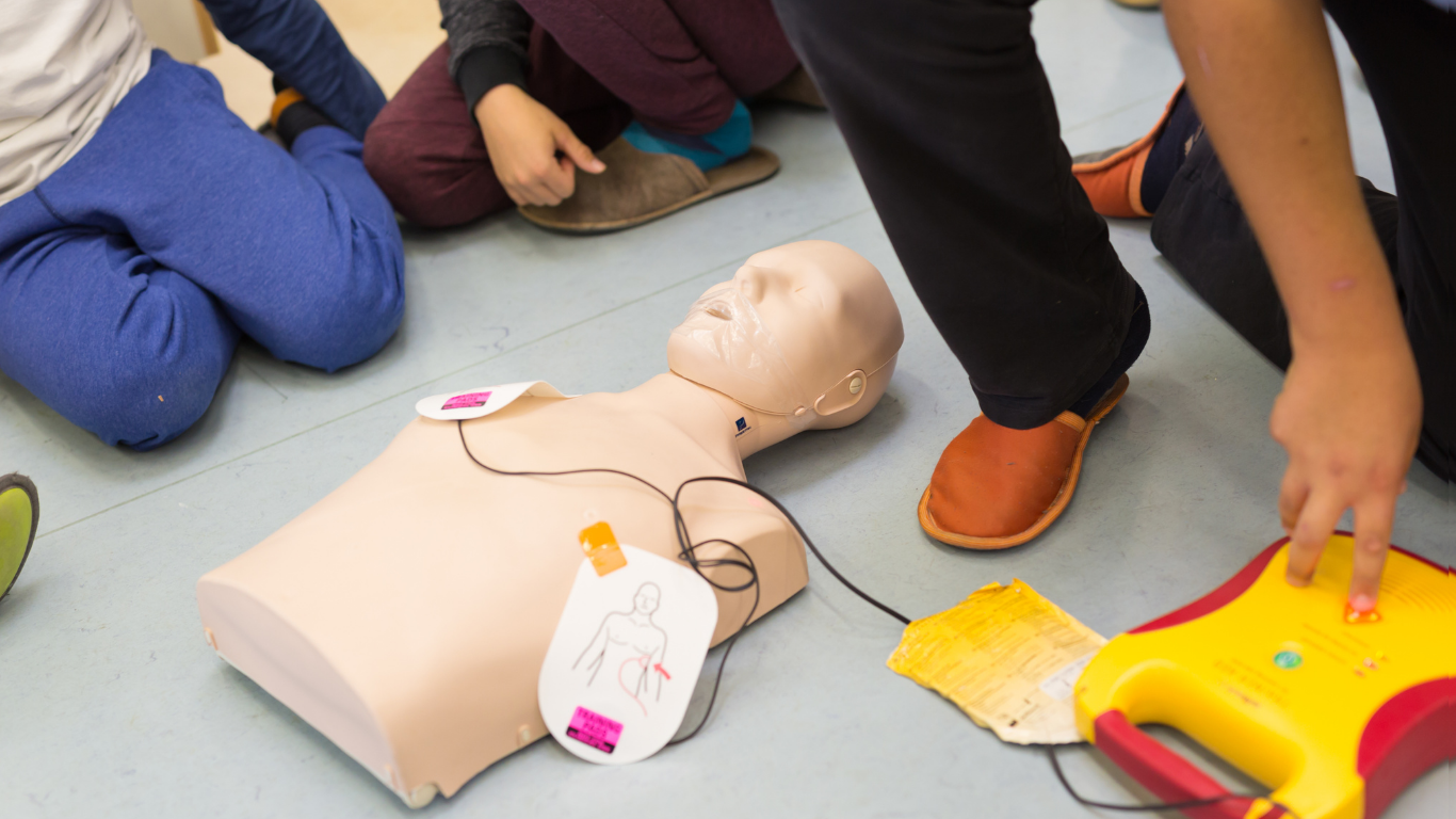 A group of children are kneeling around a mannequin with a defibrillator attached to it.