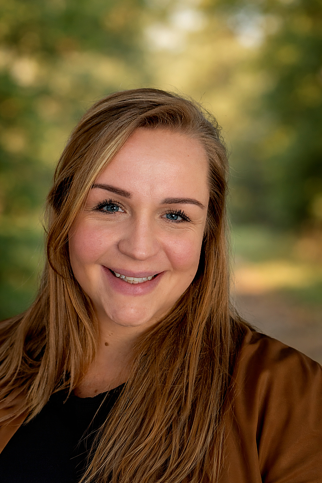 Een lachend persoon met krullend bruin haar, gekleed in een rode top en meerdere kettingen, tegen een lichte achtergrond.