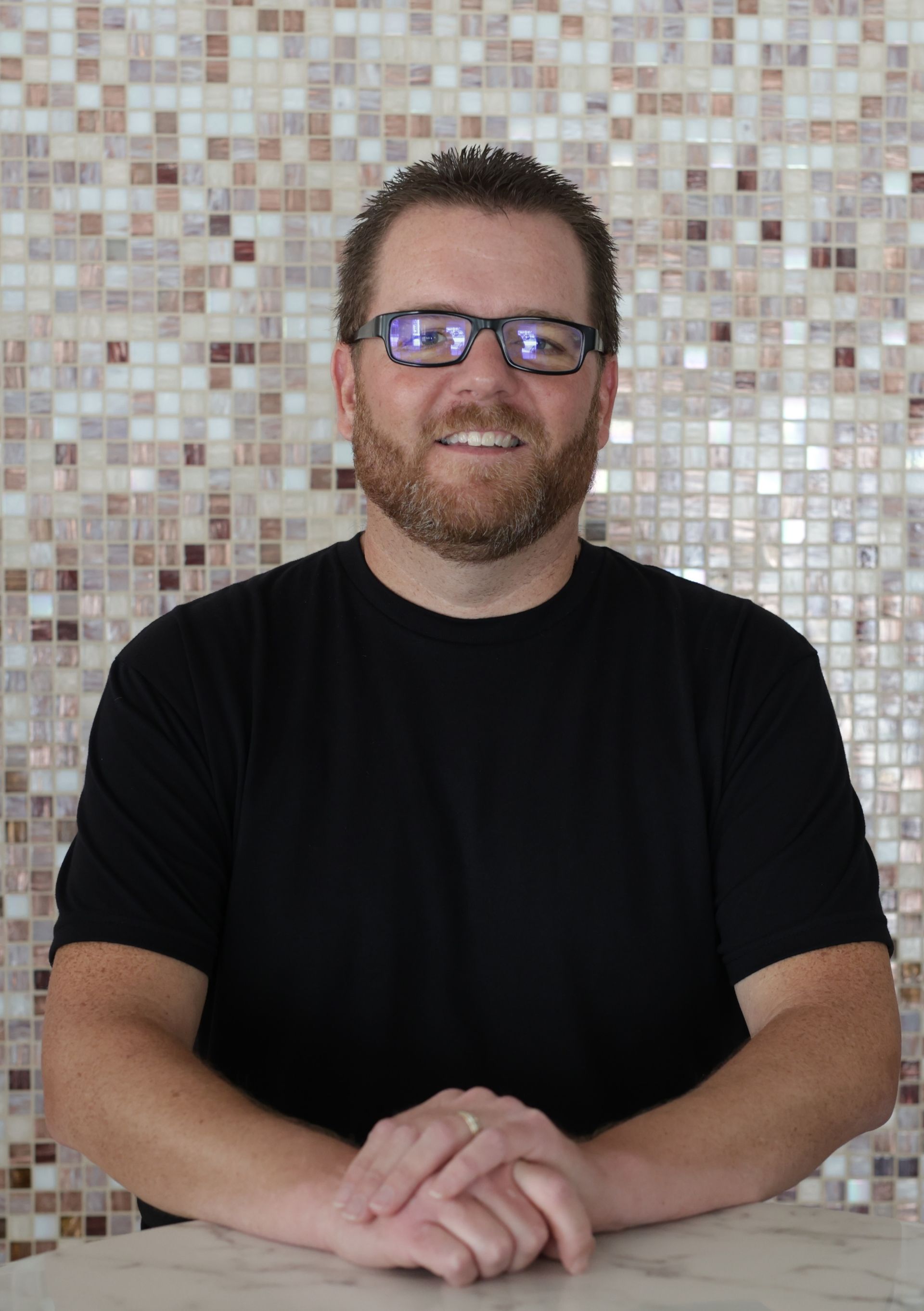 Man with dark hair and a black shirt, smiling, arms crossed. Mosaic tile background.