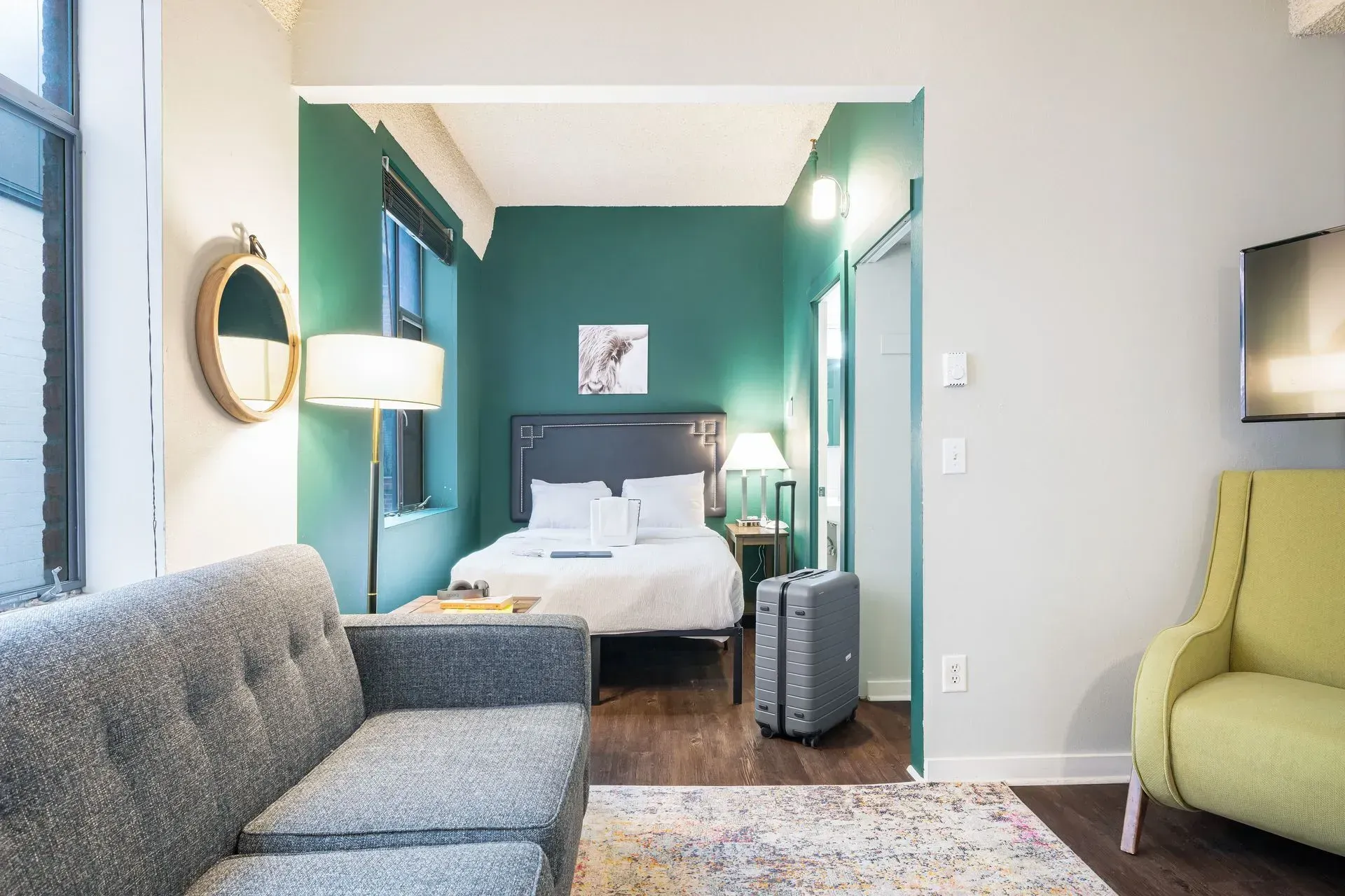 Small apartment interior: green bedroom visible from the living area with gray sofa, yellow chair, luggage, and TV.