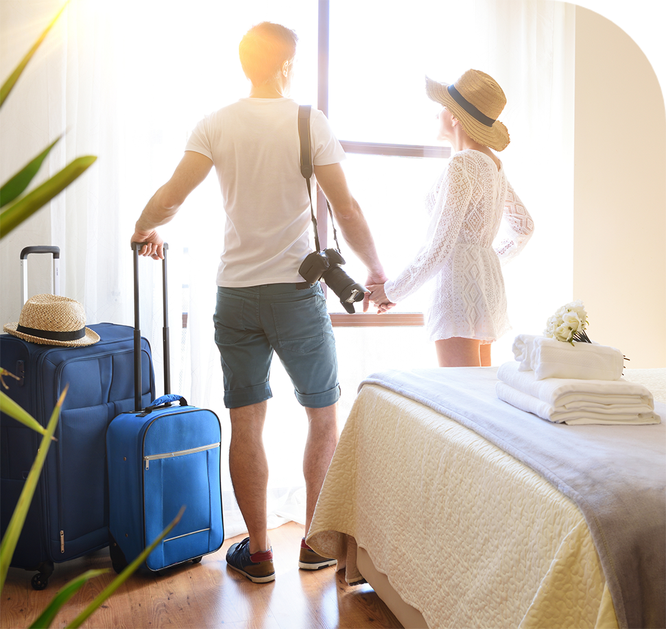 Couple holding hands, looking out a window in a hotel room, luggage beside them.