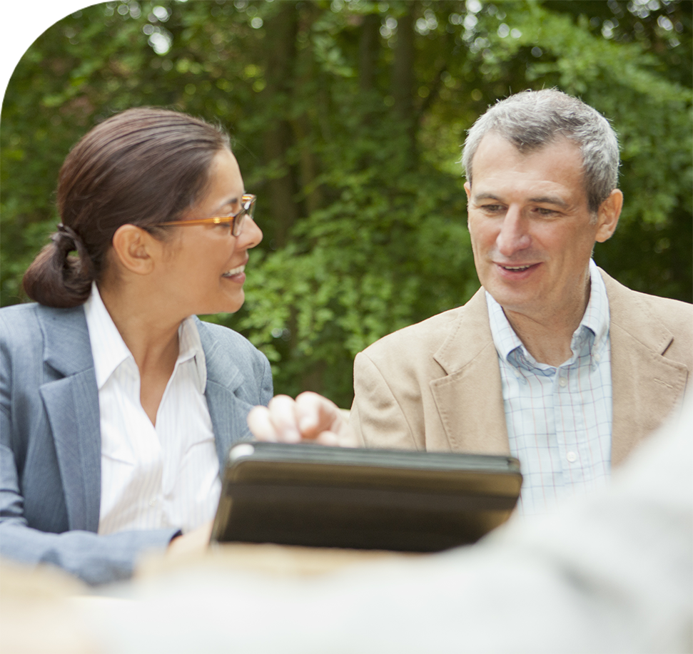 Woman and man looking at a tablet, smiling. Outdoors, wearing business attire, trees in the background.