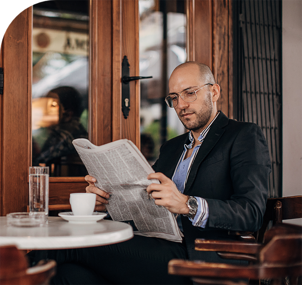 Man in suit reading newspaper at outdoor cafe table.