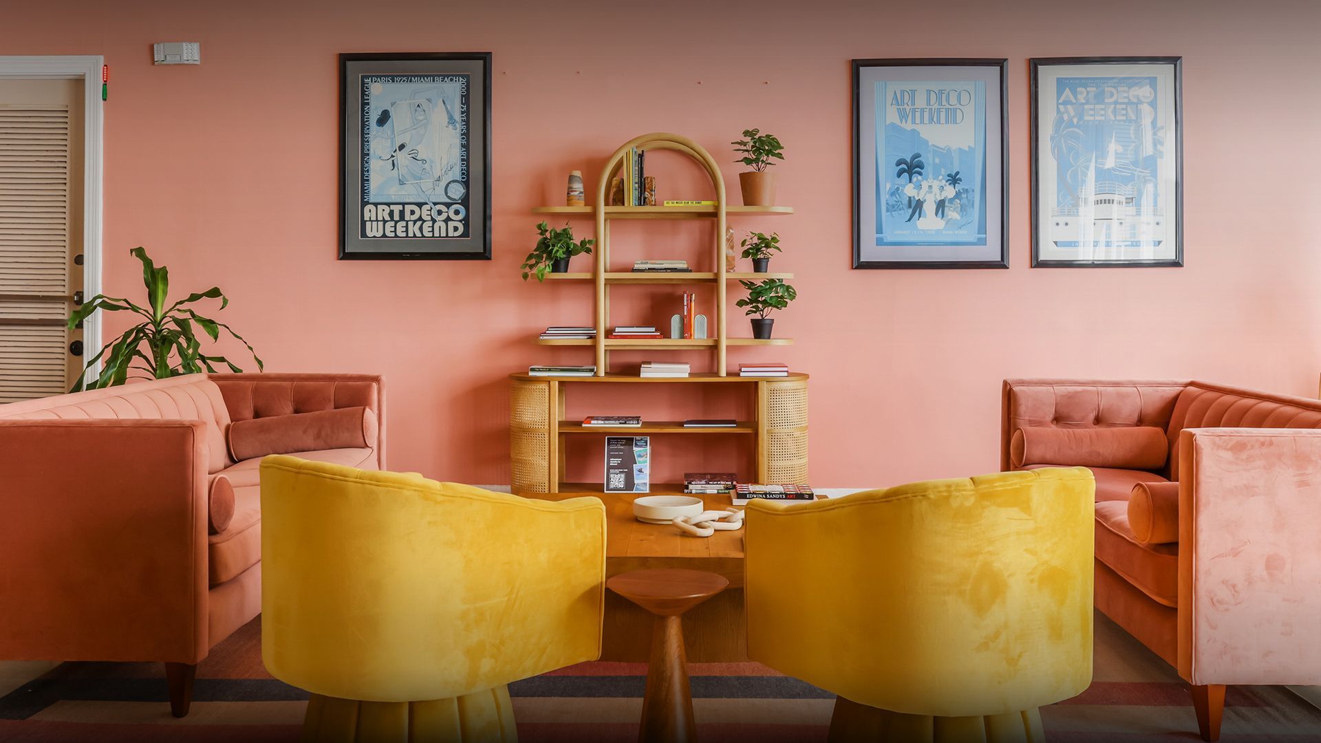 Pink-walled room with velvet pink sofas, yellow chairs, wood shelf, and framed art.