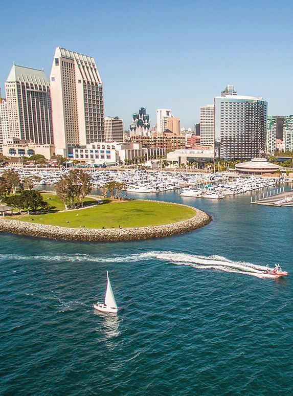 Sailboat on water, cityscape backdrop. San Diego harbor with buildings and marina. Clear blue sky.