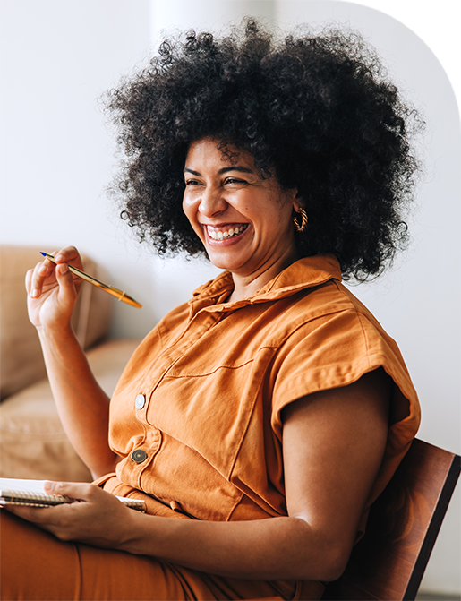 Woman with large afro smiles, holding pen and notebook in terracotta-colored outfit.