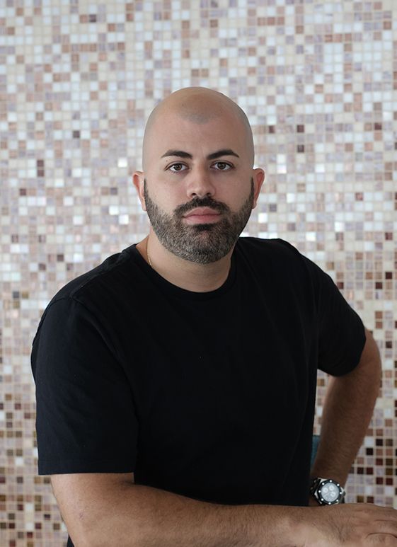 Bald man with a full beard, black shirt, posing in front of a mosaic tile wall.