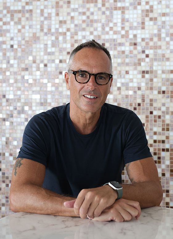 Man with glasses and navy shirt sitting at a table, arms crossed. Background has patterned tile.