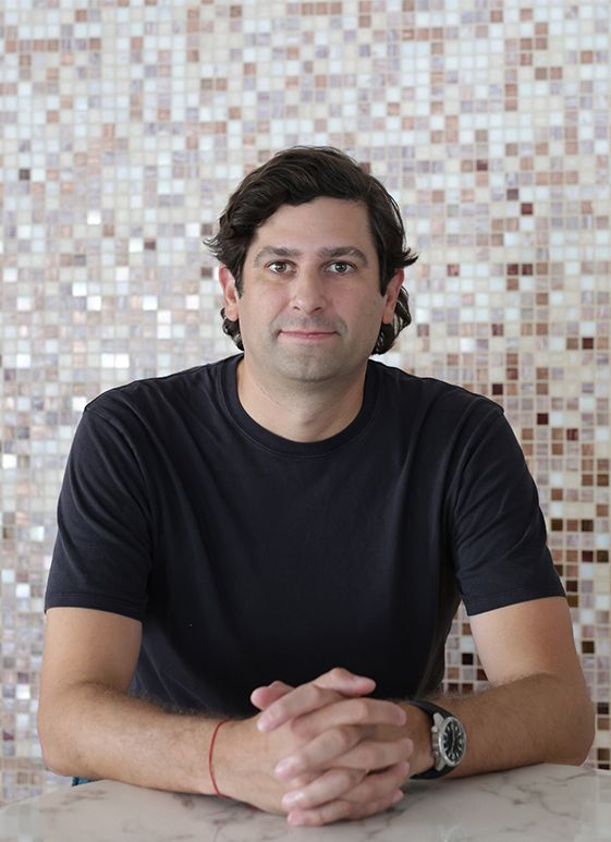 Man with dark hair and blue shirt seated, arms crossed, in front of mosaic tile wall.