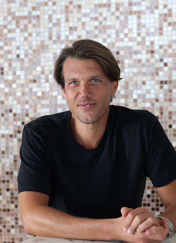 Man with light brown hair in black shirt, smiling with hands clasped, against a mosaic tile wall.