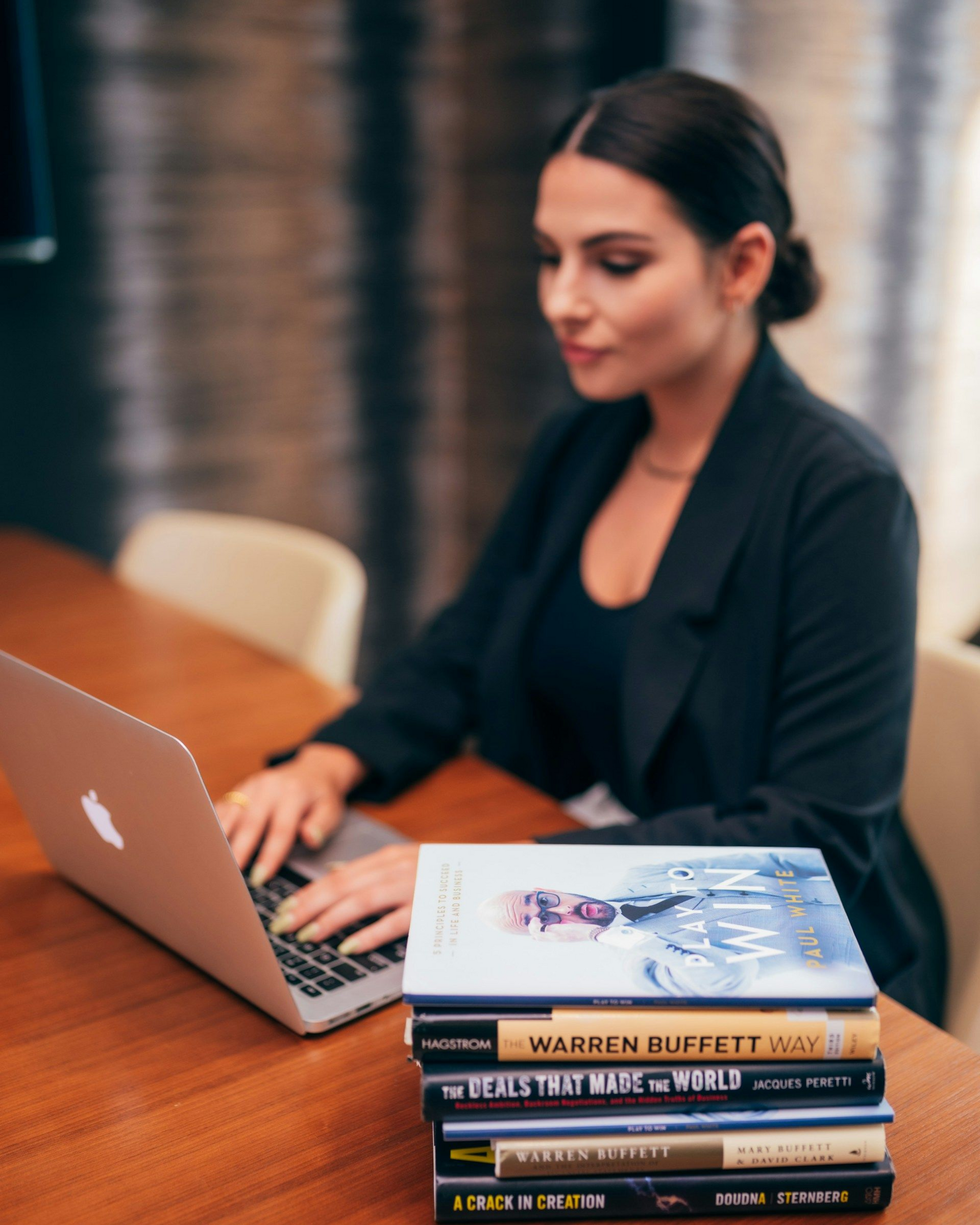Woman in blazer using a laptop, stack of books on desk.