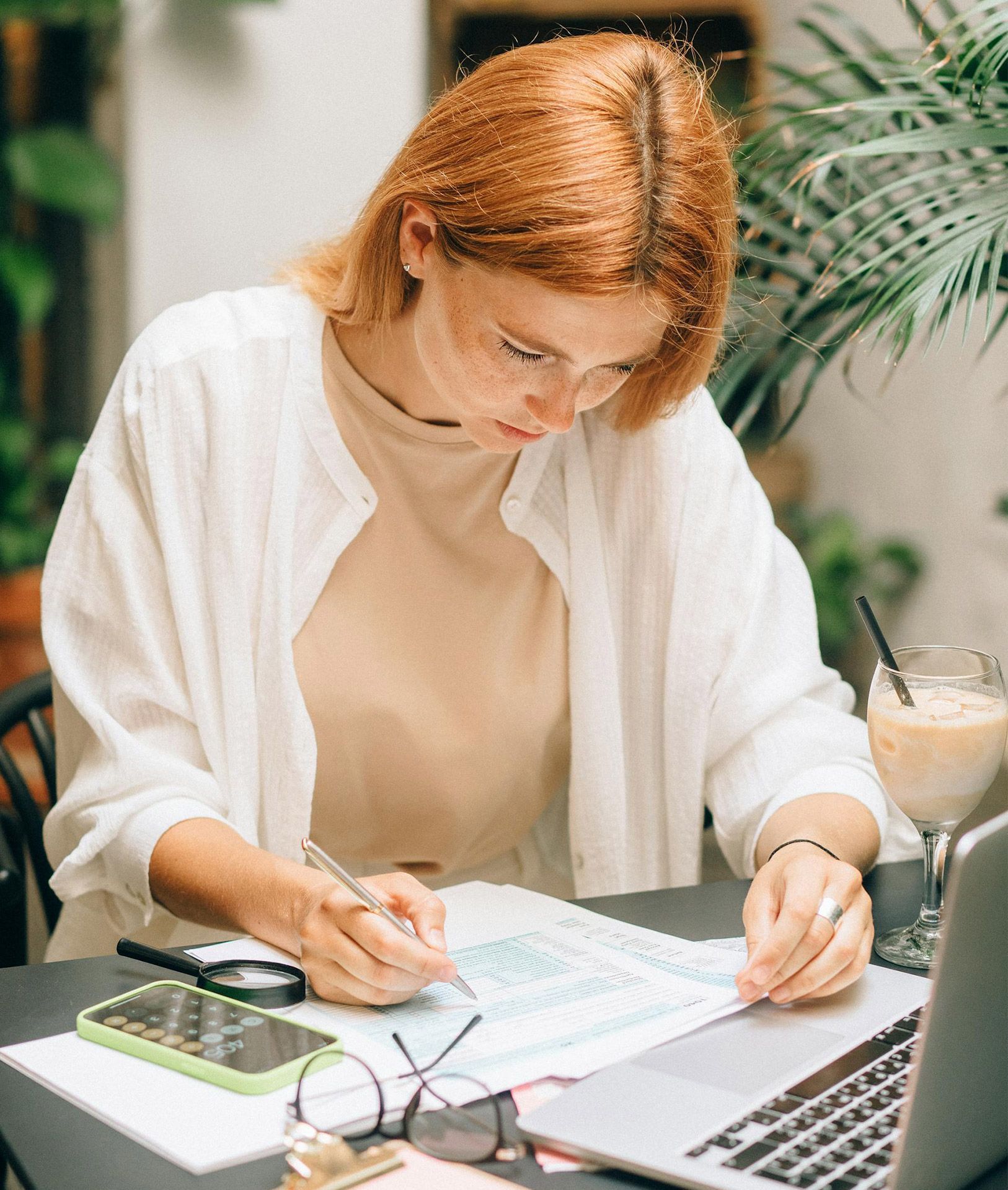 Woman writing at a table, looking at papers. Laptop, drink, and phone present.