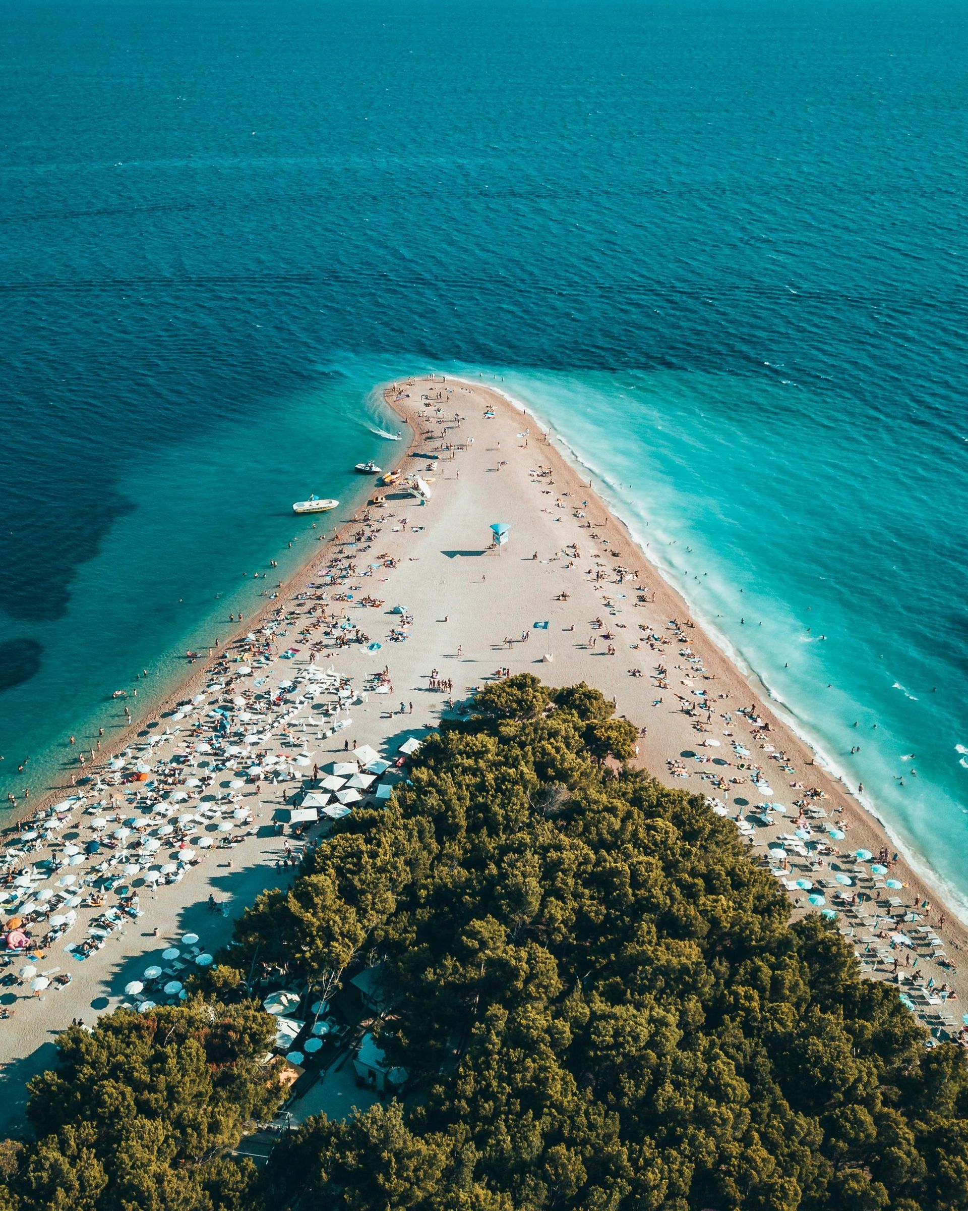 A crowded, sandy beach extends into turquoise waters, with a forest at the base.