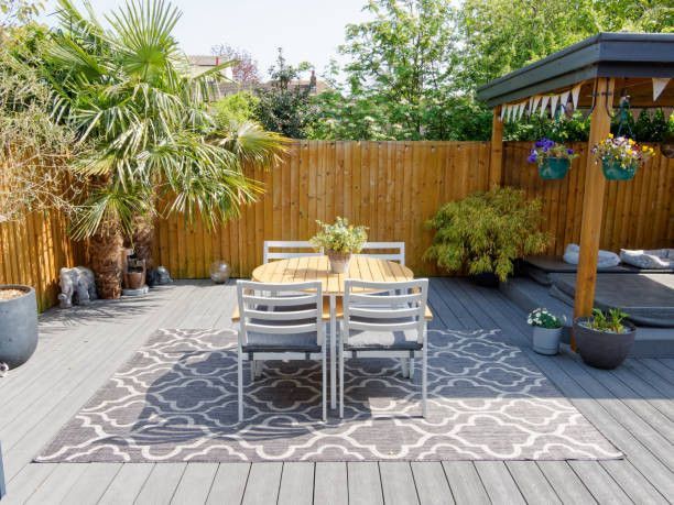 Backyard patio with dining table, rug, and plants against wooden fence.
