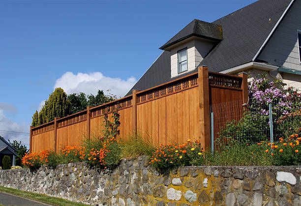 Brown wooden fence atop a stone wall, next to a house with a gray roof and blue sky.