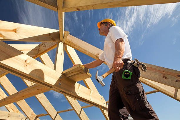 Carpenter hammering wood on a roof frame under a blue sky.
