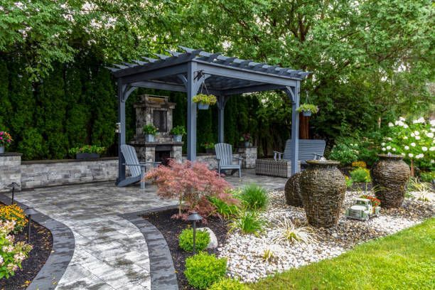 Gray pergola with stone fireplace, seating, and flowerbeds on a patio.