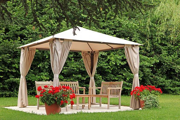 Gazebo with beige curtains, wooden furniture, and red flowers in a green garden setting.