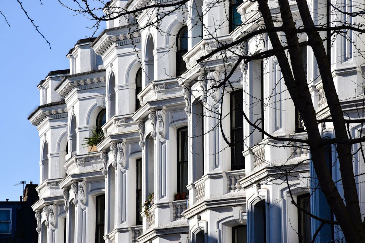 White ornate buildings with arched windows against a blue sky, tree branches frame the right side.