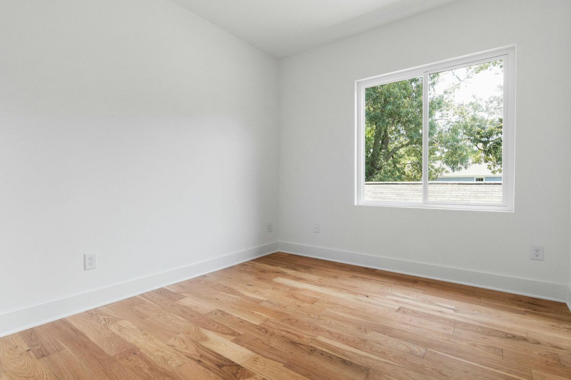Empty white-walled room with wooden floor and window overlooking greenery.