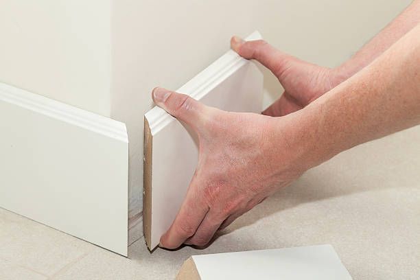 Hands fitting a white baseboard molding piece into a corner of a room.