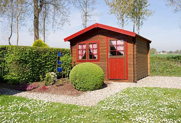 Brown wooden shed with red trim in a garden, fronted by a green bush, gravel, and flowers.