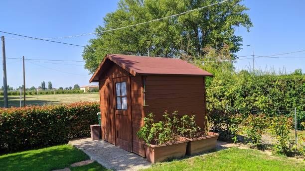 Brown wooden shed with a red roof in a sunny backyard with green grass and bushes.