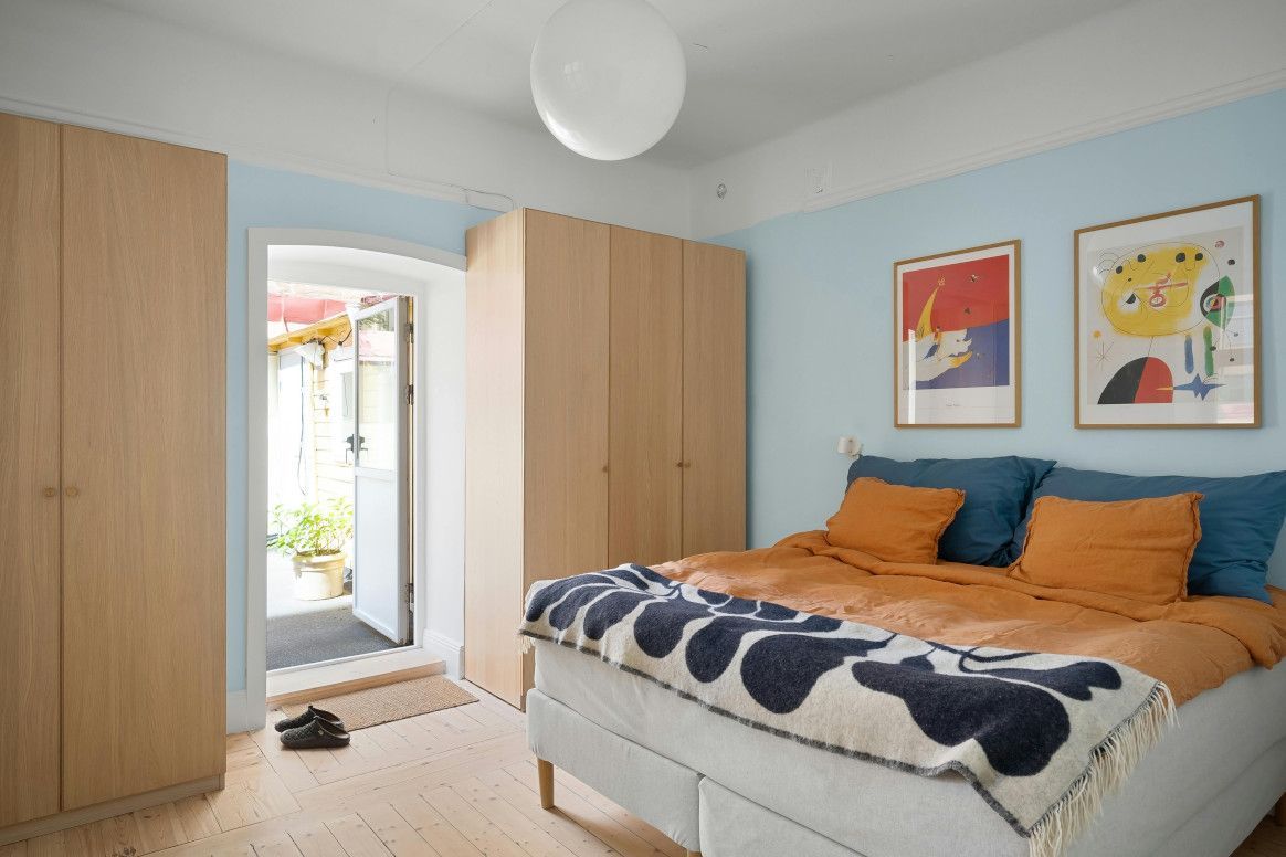Bedroom with light blue walls, bed with orange and blue pillows, wooden wardrobes, and two framed colorful art prints.