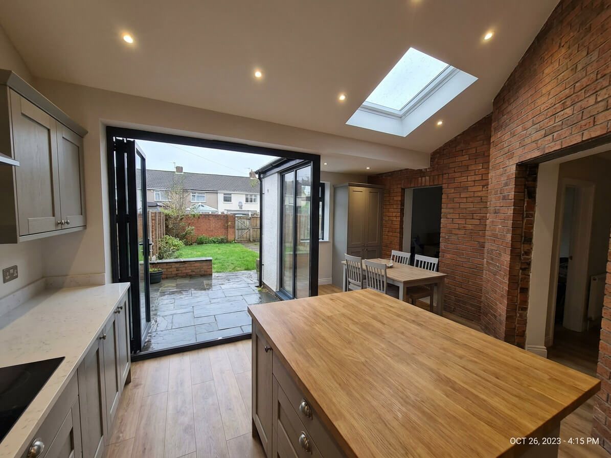 Kitchen with bi-fold doors open to a garden, light gray cabinets, wood island, and exposed brick wall.