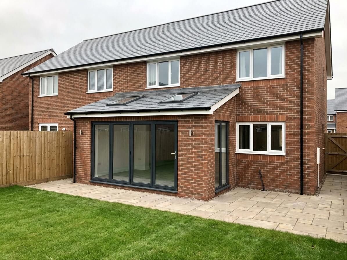 Red brick two-story house with gray roof, an extension with large glass doors, and a patio, with a green lawn.