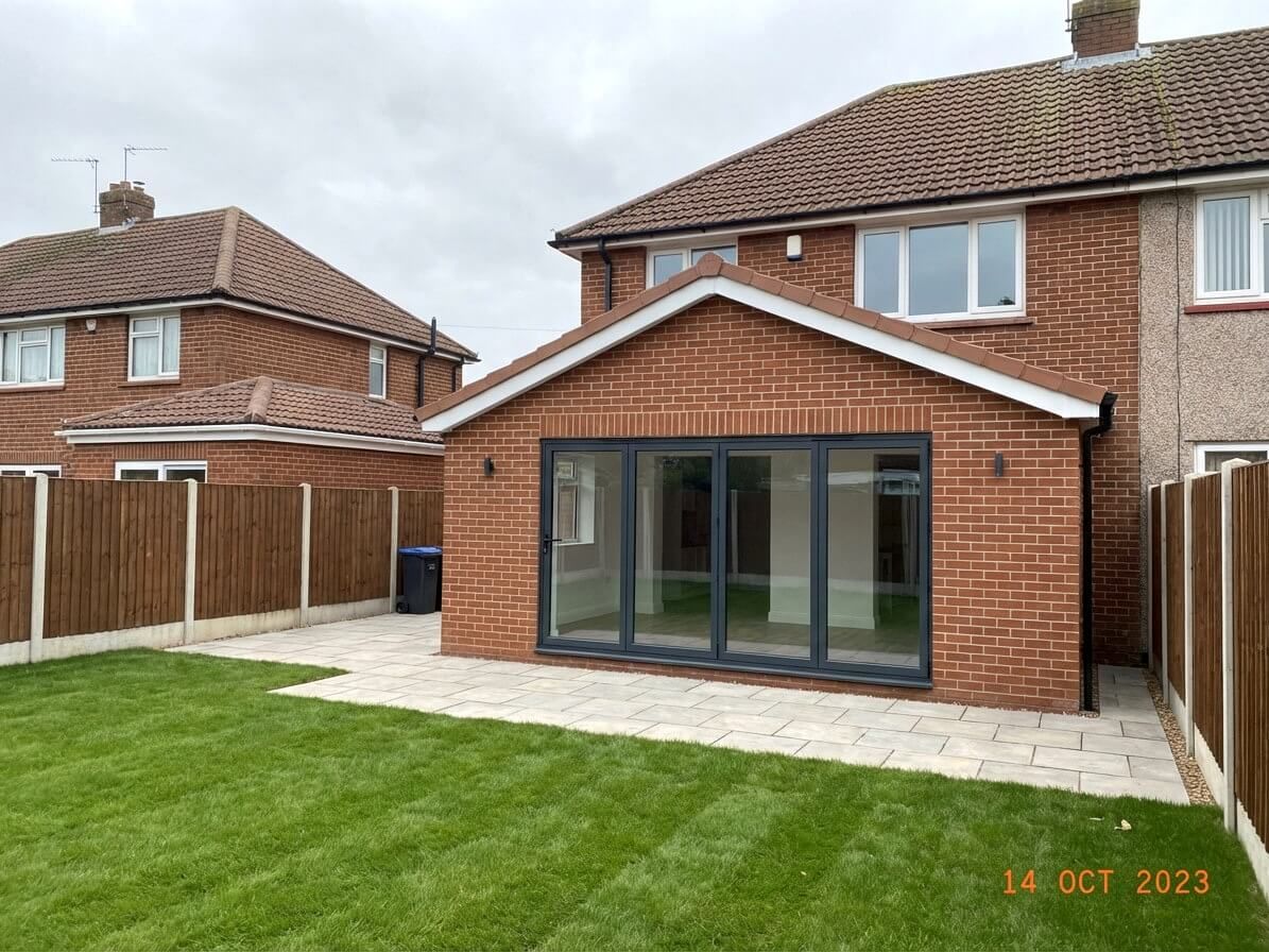 Brick house with new extension, glass doors, and patio in a grassy backyard.