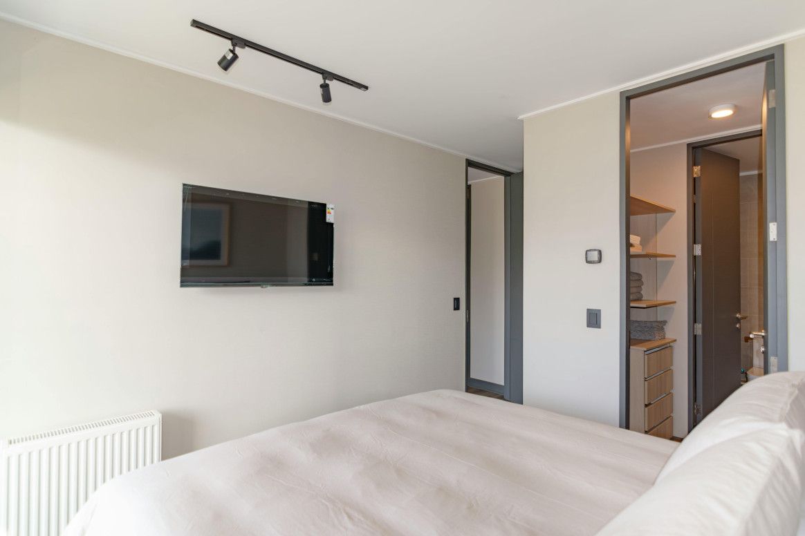 Bedroom with bed, wall-mounted TV, doorway to a closet, and a radiator. Beige walls, neutral tones.
