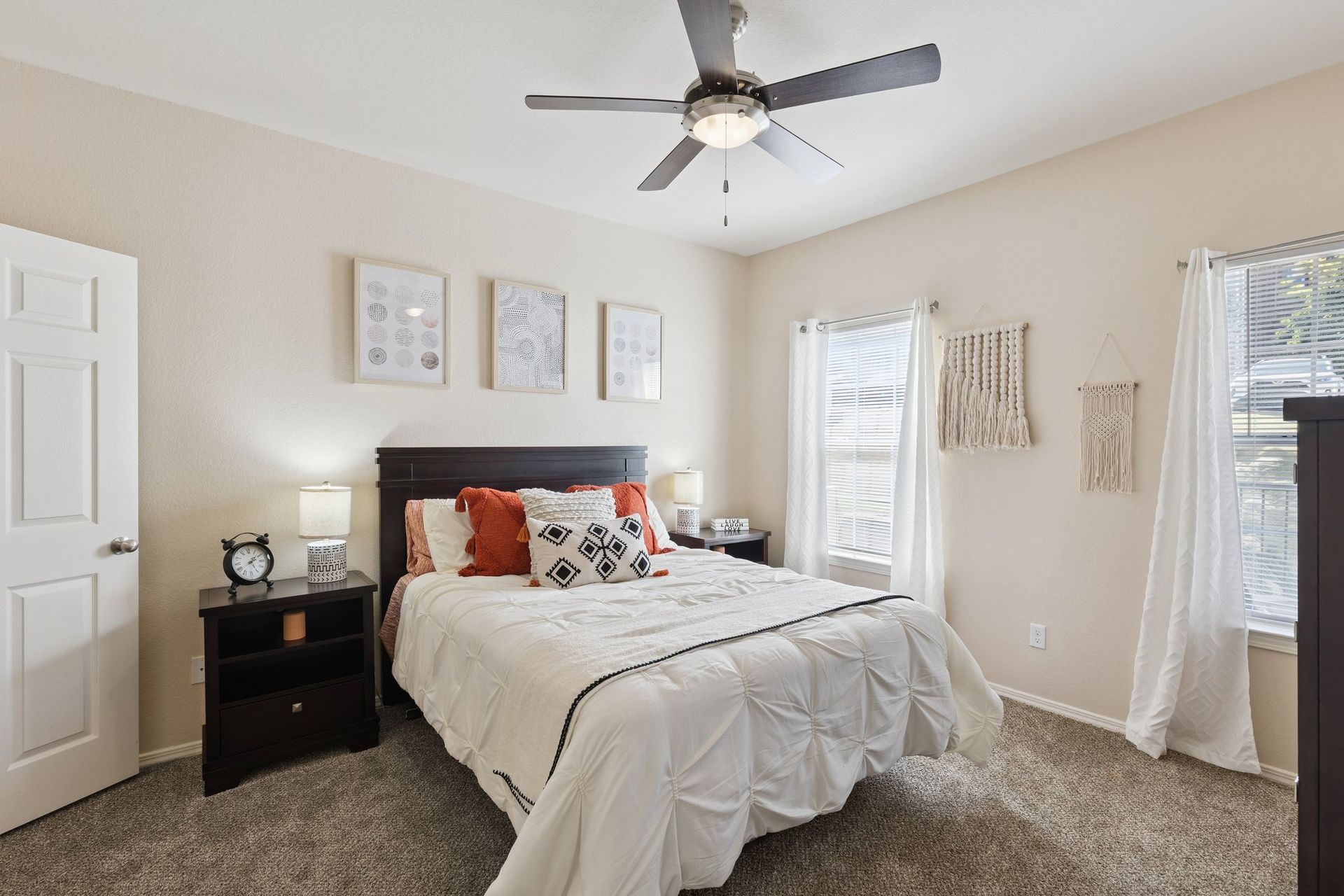 Bedroom with a bed, nightstands, wall art, windows with white curtains, and a ceiling fan.