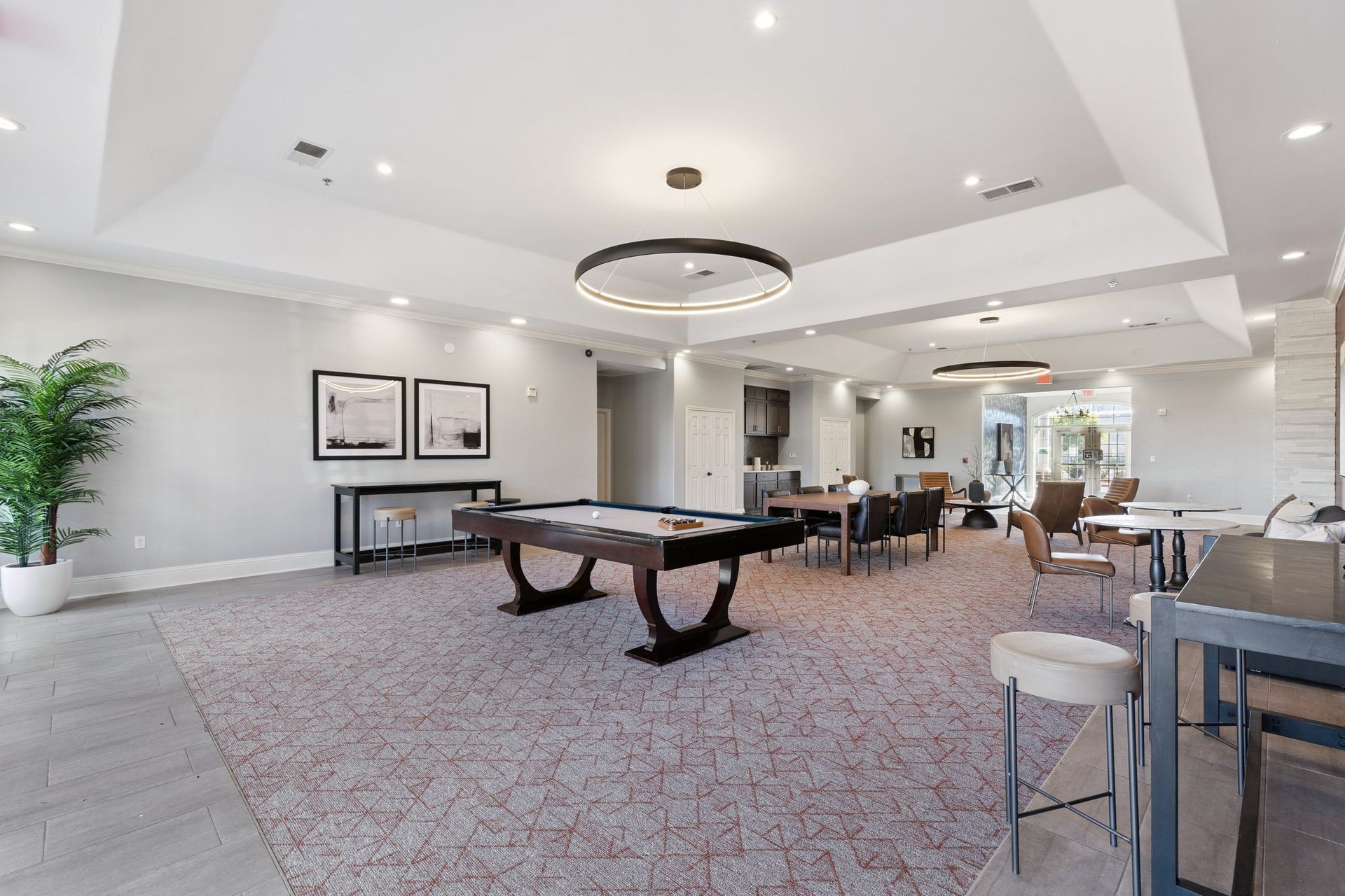 Spacious rec room with pool table, dining area, and seating. Neutral tones, overhead lighting, and potted palm.