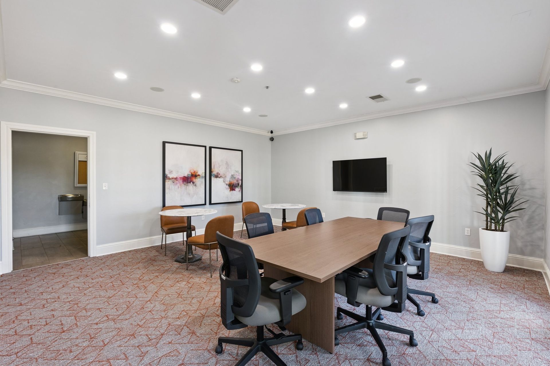 Meeting room with a large oval table, chairs, a TV, artwork, and a potted plant.