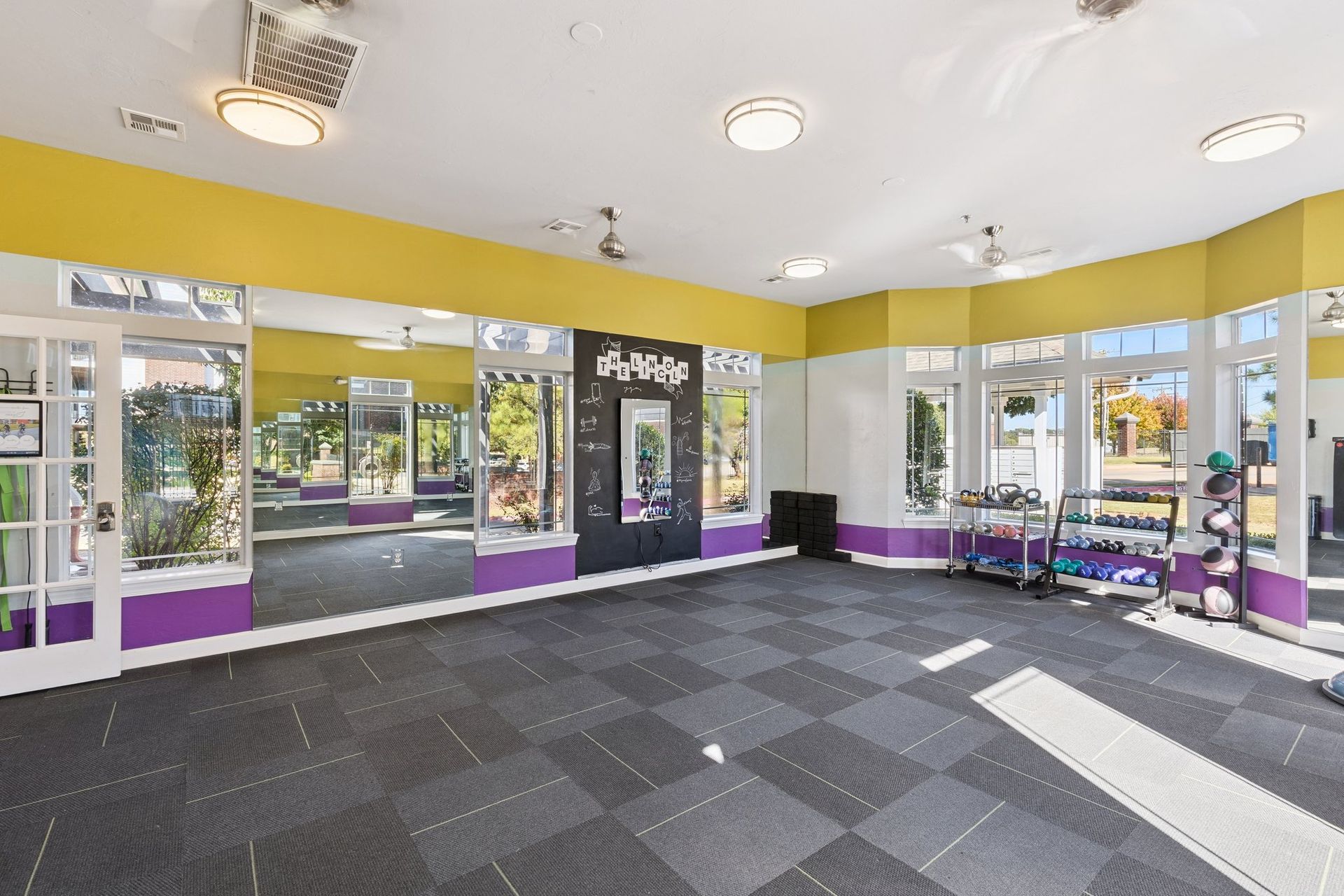 Gym interior with mirrors, exercise equipment, purple/yellow/white walls, and checkered floor.